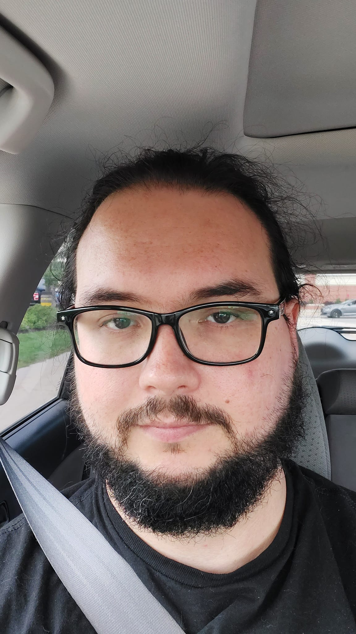 A man with long dark hair tied back sits in the passanger seat of a car